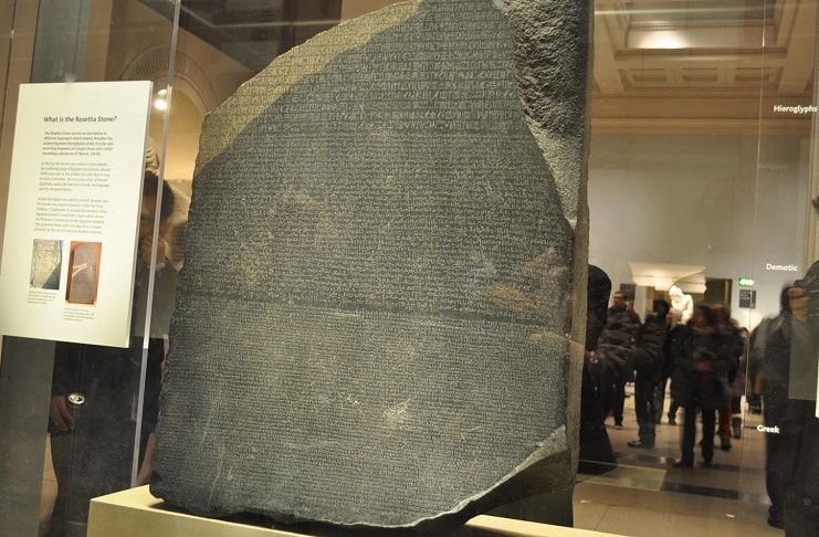 "A photograph of the Rosetta Stone on display at the British Museum, London. The dark granodiorite slab is covered in three scripts: Ancient Egyptian hieroglyphs, Demotic, and Ancient Greek."